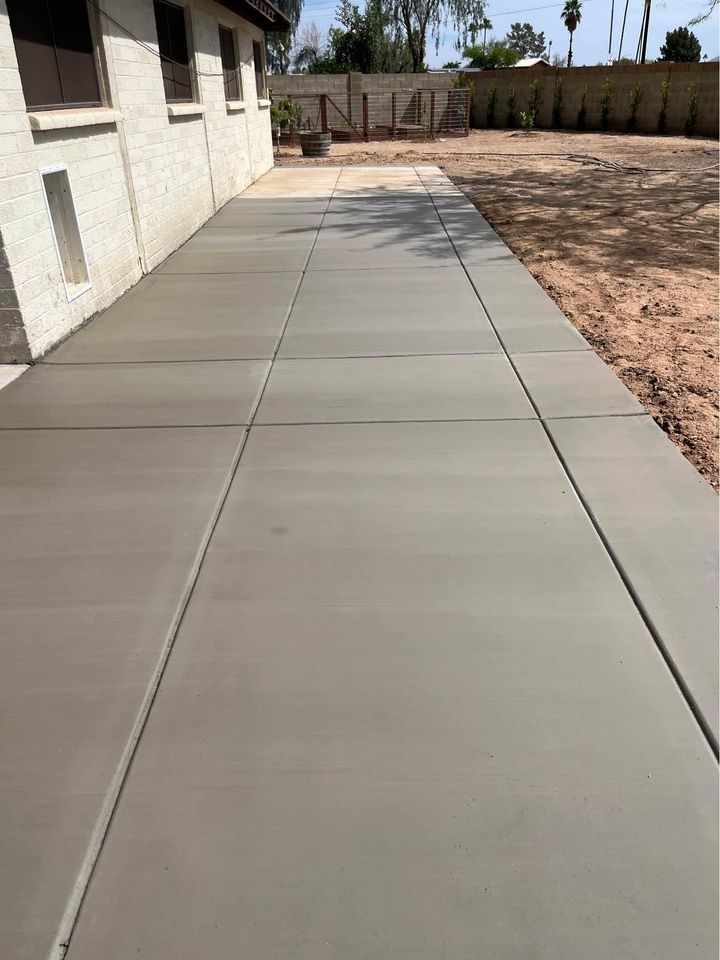 A freshly poured, smooth concrete sidewalk by a house in Riverside, CA, extending towards a fenced area in the background. The ground adjacent to the sidewalk is bare dirt, with trees and a brick wall in the distance—crafted by expert concrete contractors offering top-notch concrete services.