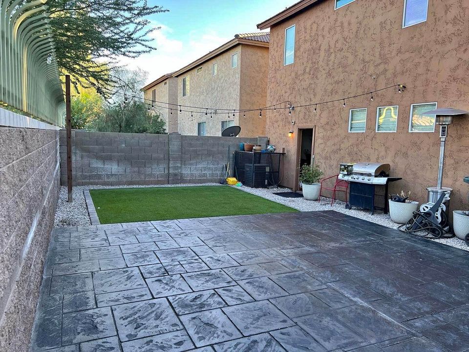 A backyard oasis with a patio showcasing dark stamped concrete, expertly crafted by skilled concrete contractors. It features a small patch of artificial grass and a cozy sitting area with a barbecue grill and string lights. Enclosed by a brick wall, two-story houses add to the backdrop charm.