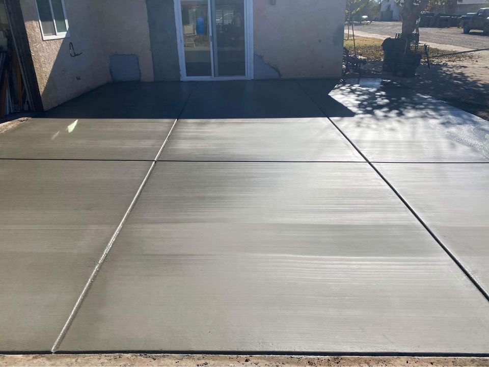 A freshly poured concrete patio with a smooth surface, consisting of four large rectangular sections, enhances the outdoor living space in front of a Riverside CA home entrance. A tree elegantly casts its shadow on the right side, completing this serene oasis.