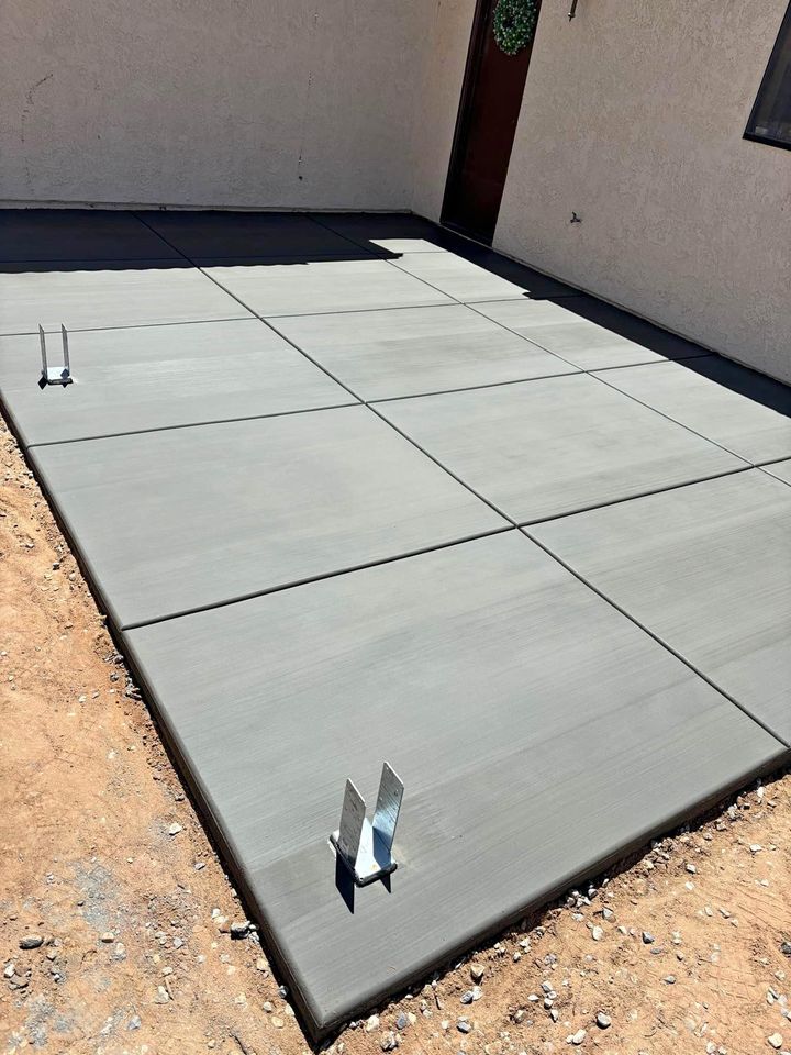 A freshly poured concrete patio with a grid pattern, showcasing the precision of concrete contractors. Visible saw cuts divide it into square sections. The area is surrounded by dirt, with metal stakes near the edges. A building wall looms in the background, framing this fine example of concrete construction.