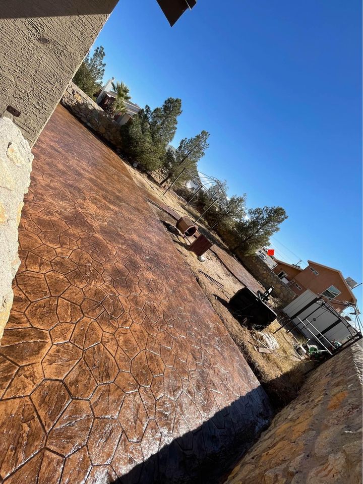 A backyard in Riverside, CA, features a newly installed textured brown stone patio. Concrete services expertly crafted the area, surrounded by a stone wall with trees and a house in the background under the clear blue sky.