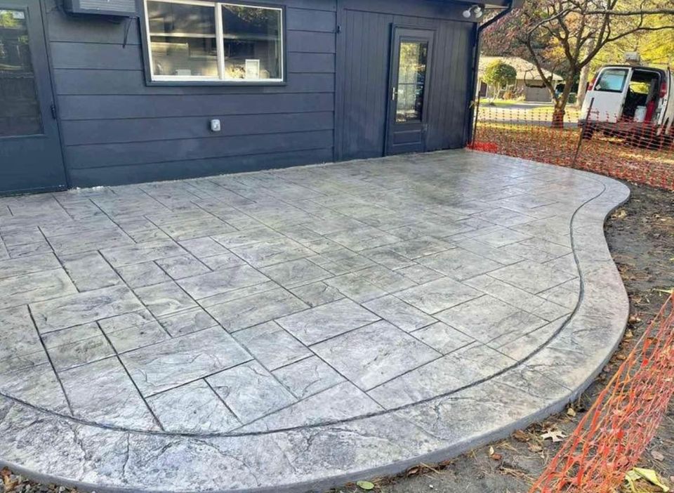 A newly installed concrete patio with a geometric pattern and smooth edges enhances the outdoor living space adjoining a dark-colored building. There are orange safety barriers and a white van in the background, while trees and fallen leaves subtly accentuate the area.