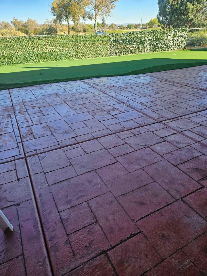 A patio with red stone tiles, crafted by expert concrete construction, glistens wet from rain. It is bordered by a hedge and offers a view of the lush, green lawn. Trees stand in the background under a clear, blue sky.