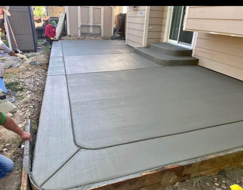 Concrete contractors in Riverside, CA meticulously smooth a freshly poured patio with intricate edges. A step leads up to the house entry, while building materials and tools are visible in the background, showcasing their expert concrete services.