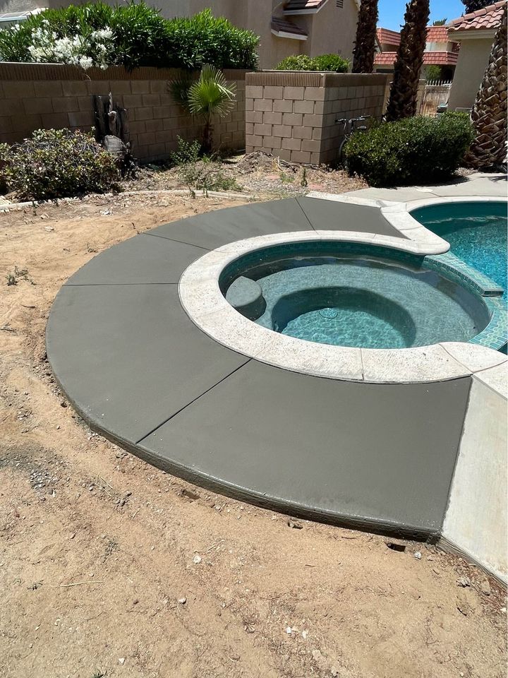 A backyard pool in Riverside, CA, features a circular spa section seamlessly integrated into a concrete pool deck. Newly laid concrete borders a bare patch of earth and a low brick wall, with lush plants and palm trees enhancing the custom design of this serene oasis.