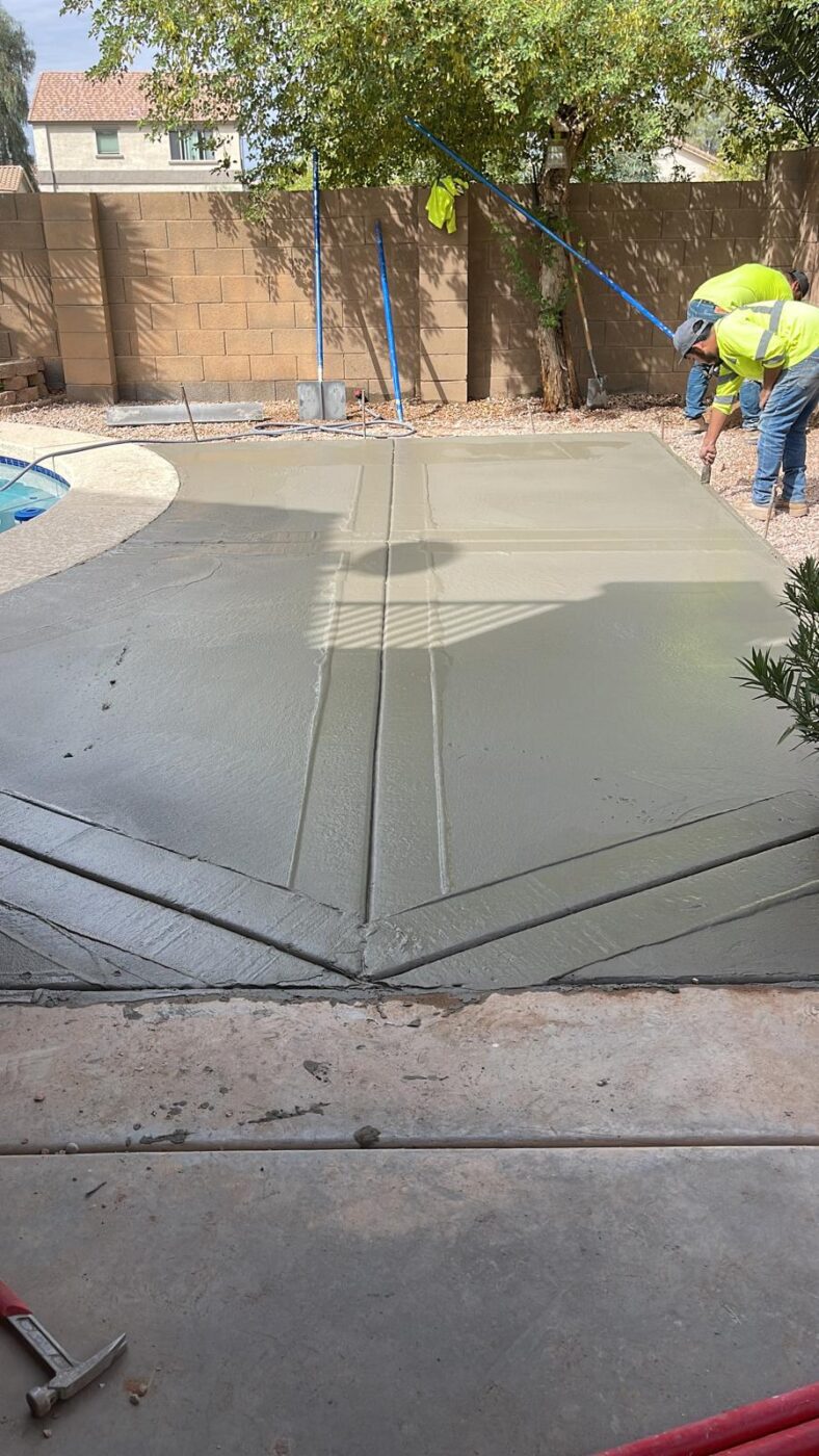 Two workers in bright yellow shirts smooth the freshly poured concrete pool deck for a backyard patio near Riverside, CA. Custom designs accentuate the area, with a tree in the background and construction tools scattered around.