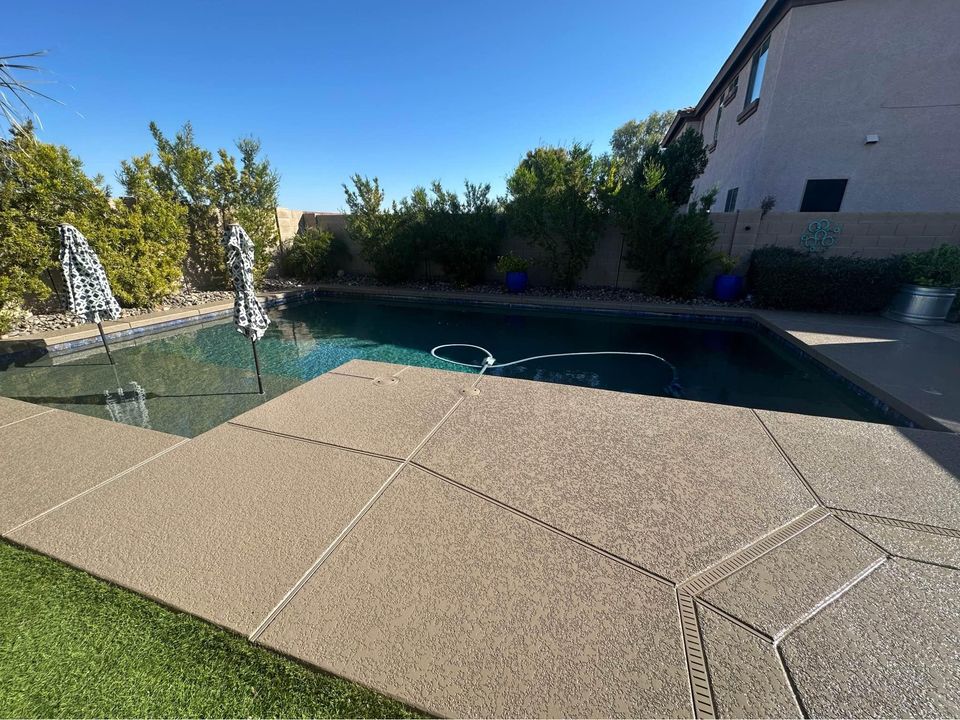 A backyard swimming pool with clear water, bordered by green bushes and trees, creates a serene escape. Two closed umbrellas stand beside the pool on a sleek concrete deck, reflecting custom designs. The surrounding area blends artificial grass and concrete, with a two-story house visible in the background.