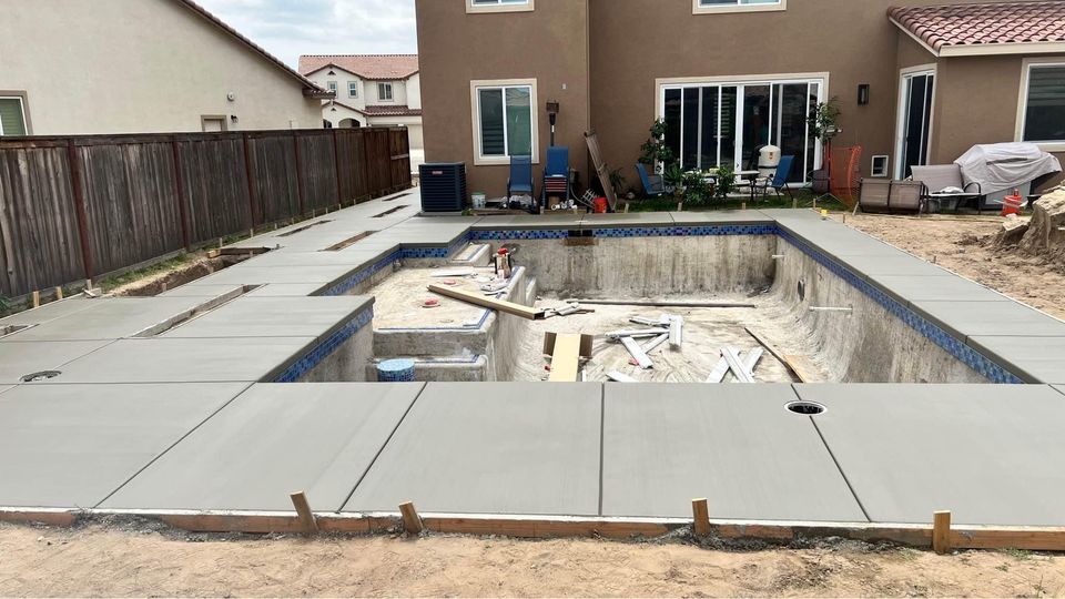 A partially constructed backyard swimming pool with new concrete decking showcases the expert work of Riverside CA's premier concrete contractors. The empty pool is surrounded by construction materials, with a brown house featuring large windows in the background and neighboring homes nearby.