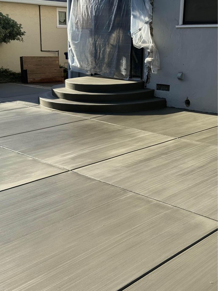 A freshly poured concrete patio, expertly crafted by skilled concrete contractors, features a set of rounded steps leading up to a door covered with protective plastic. The surface has a smooth finish with visible brush strokes, complemented by the surrounding light-colored wall.