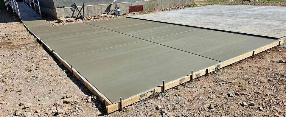 A newly poured concrete slab on a construction site in Riverside, CA, bordered by wooden forms. The surface is smooth and even, adjacent to a partially visible building with foundation contractors at work and a previously completed concrete area in the background.