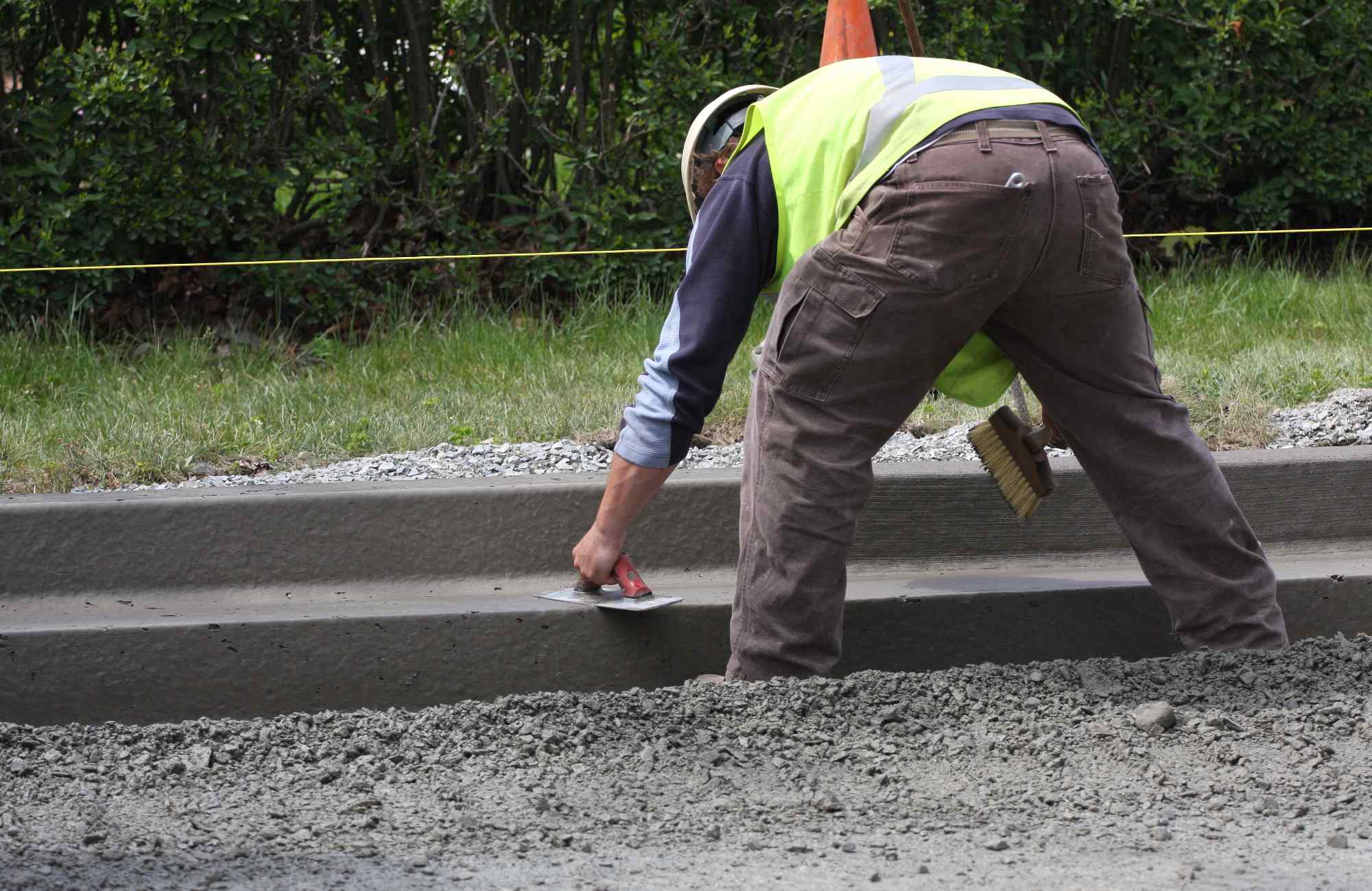 A construction worker in a neon vest smooths the surface of a concrete curb with a trowel, showcasing the precision of concrete services. Bent over and focused, their worksite is roped off with caution tape, set against a backdrop of greenery in Riverside, CA.