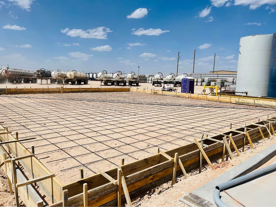 A large construction site features a rectangular foundation outlined by wood framing and a grid of steel rebar. Reliable concrete services are evident as several tanker trucks are parked in the background under a clear blue sky with scattered clouds. A portable toilet stands on the right.