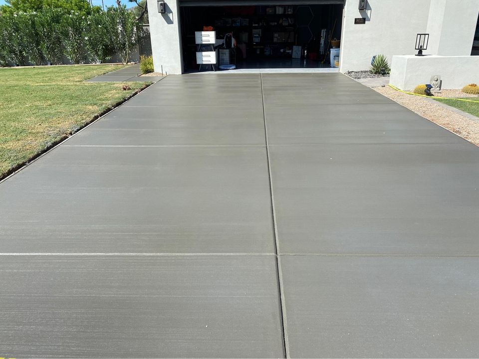 A smooth, decorative concrete driveway crafted by expert concrete contractors in Riverside, CA, leads up to an open garage filled with various items. The surrounding lawn is neatly trimmed, and there's a path to the left side of the house.