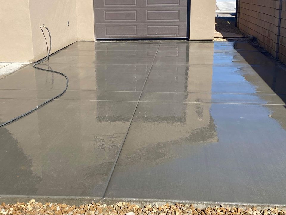 A freshly poured decorative concrete driveway stretches before a garage with its door closed. The smooth, wet surface mirrors parts of the building and sky, while a garden hose rests to the left.