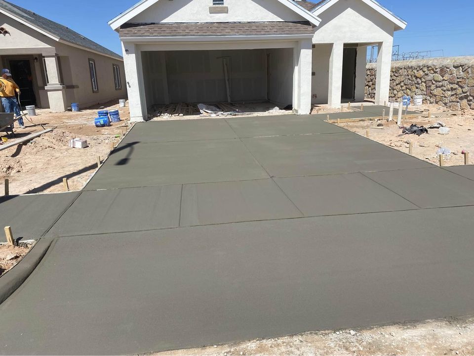 A freshly poured decorative concrete driveway in front of a two-car garage boasts a smooth and even surface. A person stands to the side as the house, under construction in Riverside, CA, displays building materials scattered around the site.