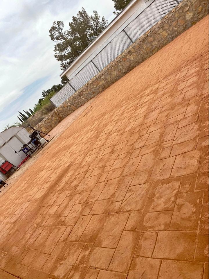 A large, empty outdoor area in Riverside, CA features a decorative concrete floor pattern, surrounded by a stone wall and a fenced building. Beneath the cloudy sky, trees are visible in the background. Some outdoor furniture is on the left side.