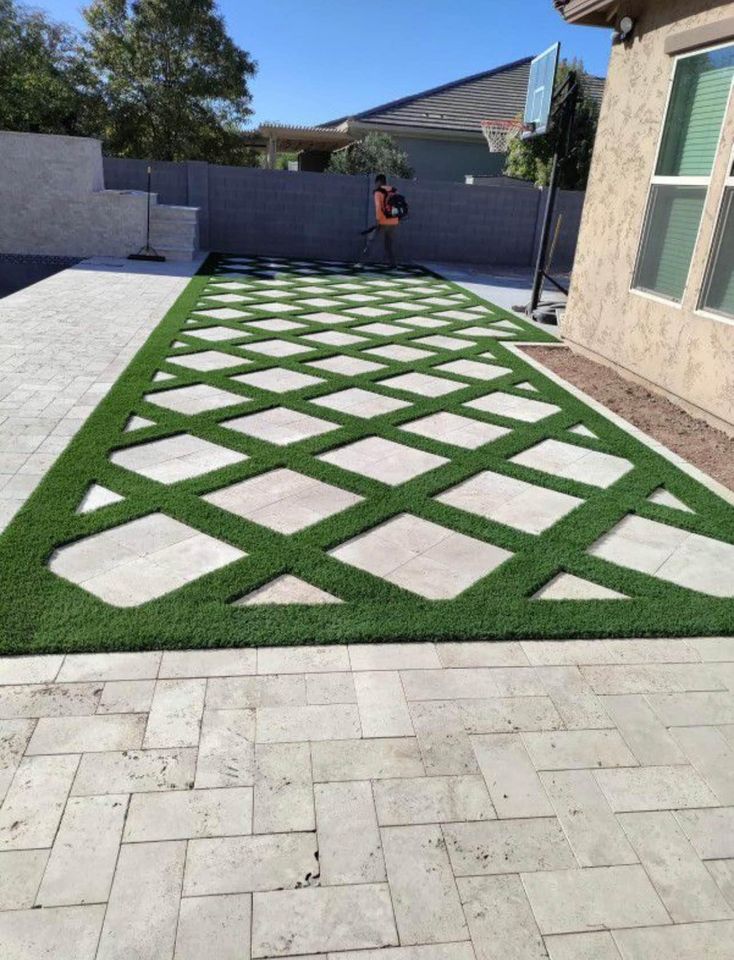 A backyard in Riverside, CA, showcases a modern geometric design with lush green grass and light stone tiles arranged in a diamond pattern. A person in the background uses a leaf blower, while concrete contractors expertly installed the basketball hoop at the far end.