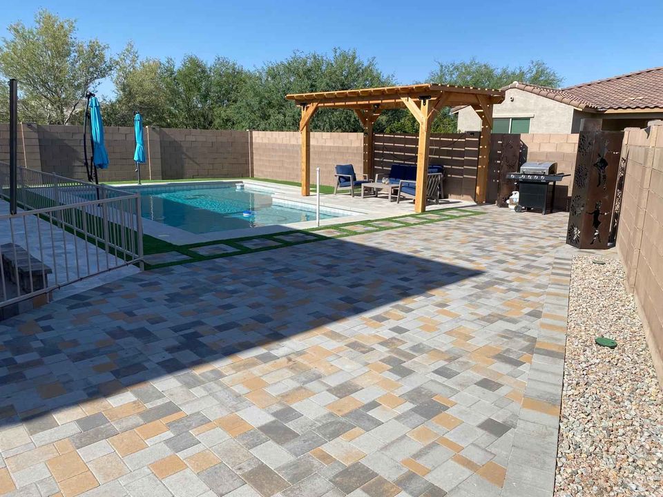 A backyard in Riverside CA with a stone patio features a pool enclosed by a white fence. This inviting outdoor living space includes a wooden pergola with patio furniture, alongside a grill and blue umbrellas. The yard is bordered by a brick wall, with trees providing background shade.