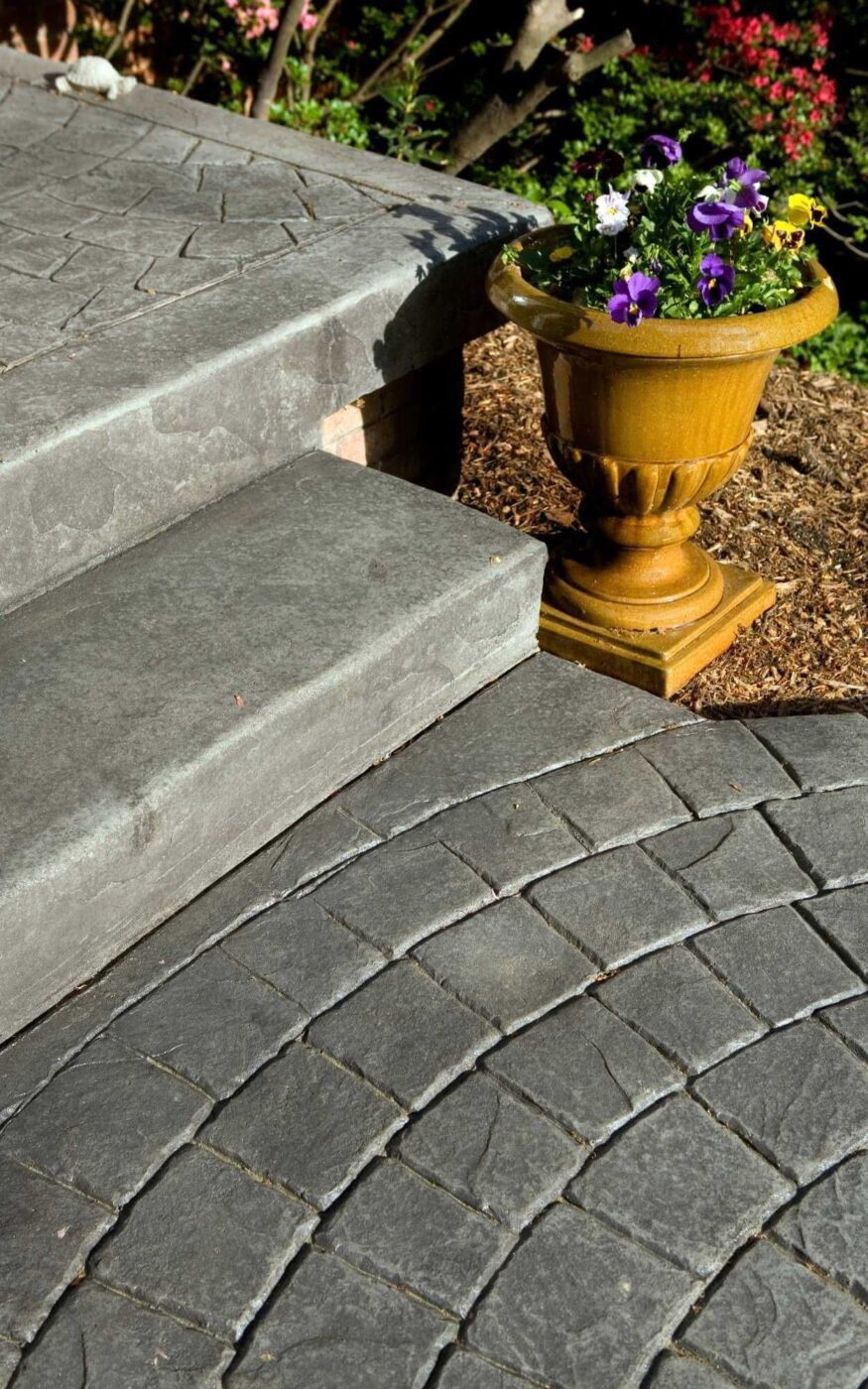 Stone steps with a curved, textured surface crafted by skilled concrete contractors lead up to a garden area. A large, yellow planter with purple and white flowers sits on the edge, surrounded by mulch and blooming plants in the background.