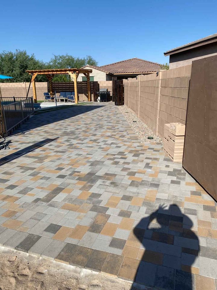 A backyard in Riverside, CA features a paved patio with multi-colored tiles extending towards a wooden pergola. There's a fence on the left and the shadow of someone snapping a picture. This outdoor living space is bordered by desert landscaping under a clear blue sky.